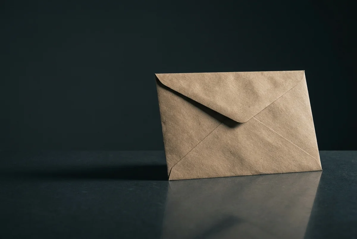 Envelope pardo lacrado sobre superfície de grafite, com sombra alongada projetada por luz rasante lateral