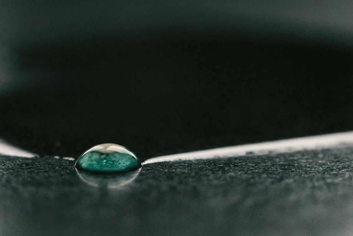 Gota de líquido azul-esverdeado sobre superfície grafite polida, com reflexo alongado sob luz rasante