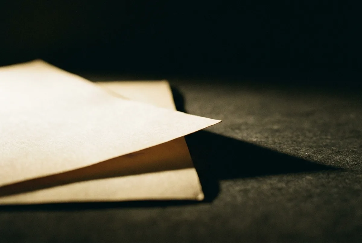 Duas folhas de papel sobrepostas sobre mesa grafite, a de cima levemente deslocada, sob luz lateral dura