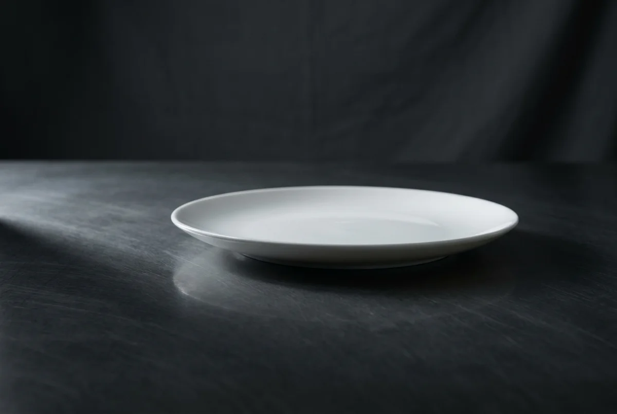 Prato de porcelana branca vazio sobre superfície grafite polida, com sombra alongada sob luz rasante lateral